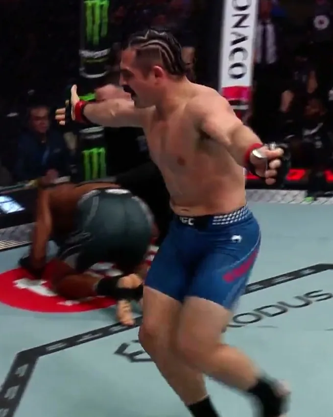 Luis Hernandez celebrating with his arms spread wide as Dumas stays down on the mat